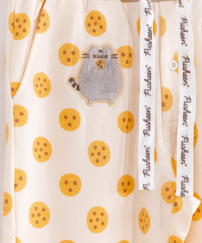 Close up of the Pusheen patch on the Pajama set pants. The patch features a fuzzy Pusheen standing upright and eating a cookie. The patch is on top of the pant's repeating cookie pattern. On the right the buttons around the groin and a silk ribbon featuring the Pusheen logo can be seen.