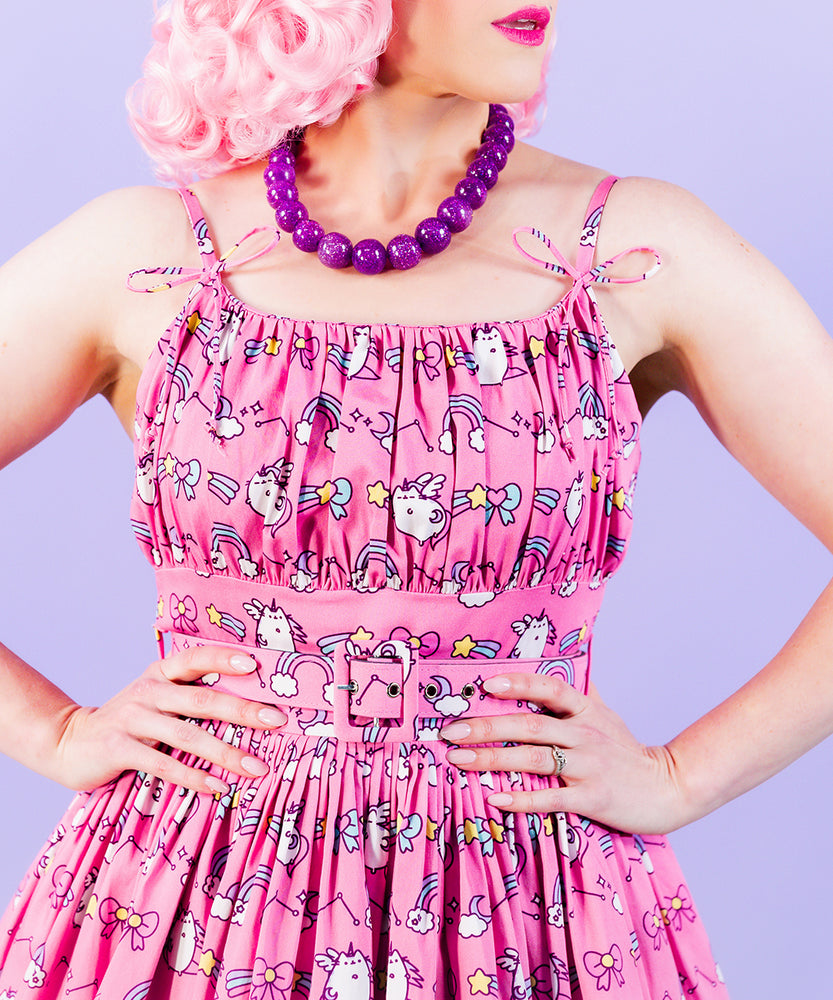 Closeup view of pleated, fitted bodice, and the string dress straps with a bow tied near the dress. The pattern on the dress are rows of a repeating string of graphics featuring Super Pusheenicorn, rainbows, and stars.  Model wears a purple necklace and has the hands placed on the waist and brings focus to the fabric covered belt with metal eyelets. 
