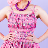 Closeup view of pleated, fitted bodice, and the string dress straps with a bow tied near the dress. The pattern on the dress are rows of a repeating string of graphics featuring Super Pusheenicorn, rainbows, and stars.  Model wears a purple necklace and has the hands placed on the waist and brings focus to the fabric covered belt with metal eyelets. 