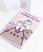 The Many Lives of Pusheen the Cat book resting at an angle on top of a white background, with white cubes above the book. 