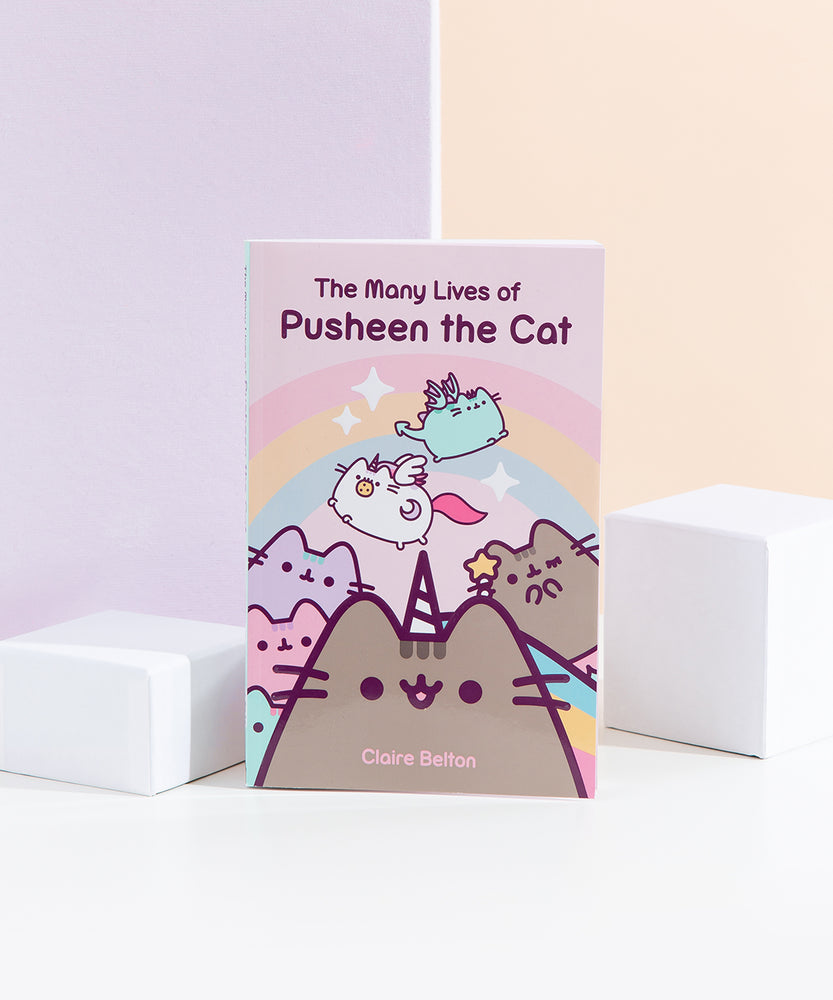 A paperback book being propped up by two white blocks in front of a pink and yellow background. The book is titled ‘The Many Lives of Pusheen the Cat’ and features Pusheenicorn in the middle, a winking Mermaid Pusheen on the right, the Pastel Pusheen on the left, Super Pusheenicorn carry a cookie and Dragonsheen flying above in front of a rainbow with sparkles. 