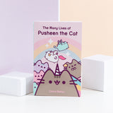 A paperback book being propped up by two white blocks in front of a pink and yellow background. The book is titled ‘The Many Lives of Pusheen the Cat’ and features Pusheenicorn in the middle, a winking Mermaid Pusheen on the right, the Pastel Pusheen on the left, Super Pusheenicorn carry a cookie and Dragonsheen flying above in front of a rainbow with sparkles. 