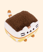 Top quarter view of the squisheen shows the fuzzy dark brown material that sits atop the square shaped plush figure. Pusheen’s tail extending off the back of the square-shaped plush.  