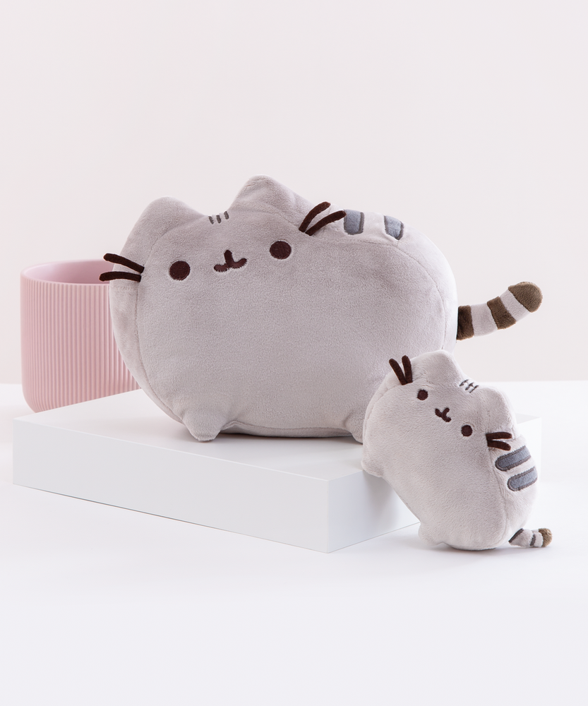The Medium Pusheen Plush on a white square platform, with a mini Pusheen plush angled in front as a size comparison for the Medium Plush. The two plush are in front of a pink ribbed pot and a light grey background.