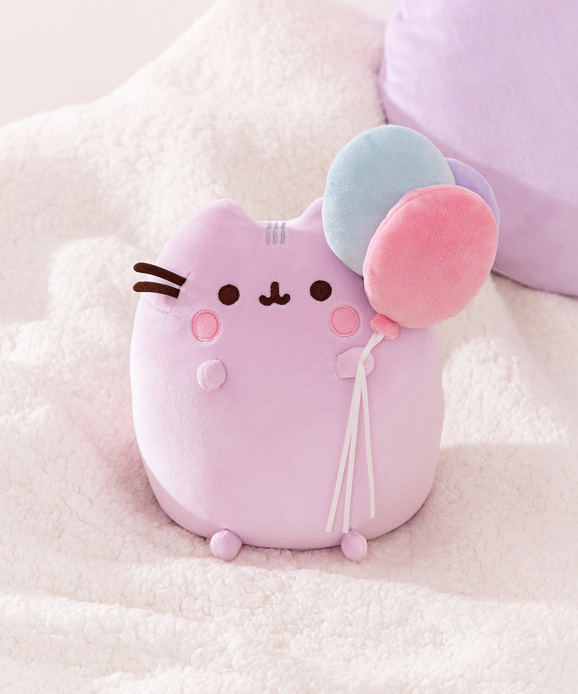 Pusheen 15th Anniversary Squisheen Plush sits up on a fluffy surface. The cat's back paws extend off the bottom of the plush while her front paws are near the embroidered facial features.