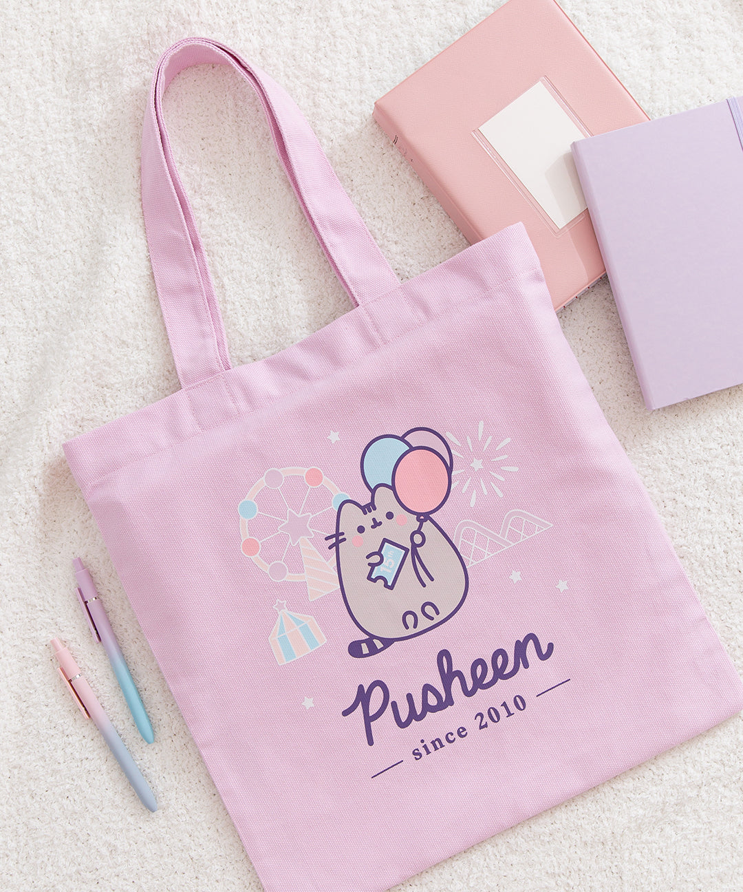 Close-up view of the printed graphic on the front of the Pusheen 15th Anniversary Tote Bag. The light grey and purple tabby cat is holding a light blue ticket while holding three pastel colored balloons. 