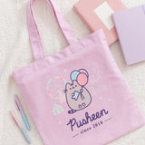 Close-up view of the printed graphic on the front of the Pusheen 15th Anniversary Tote Bag. The light grey and purple tabby cat is holding a light blue ticket while holding three pastel colored balloons. 