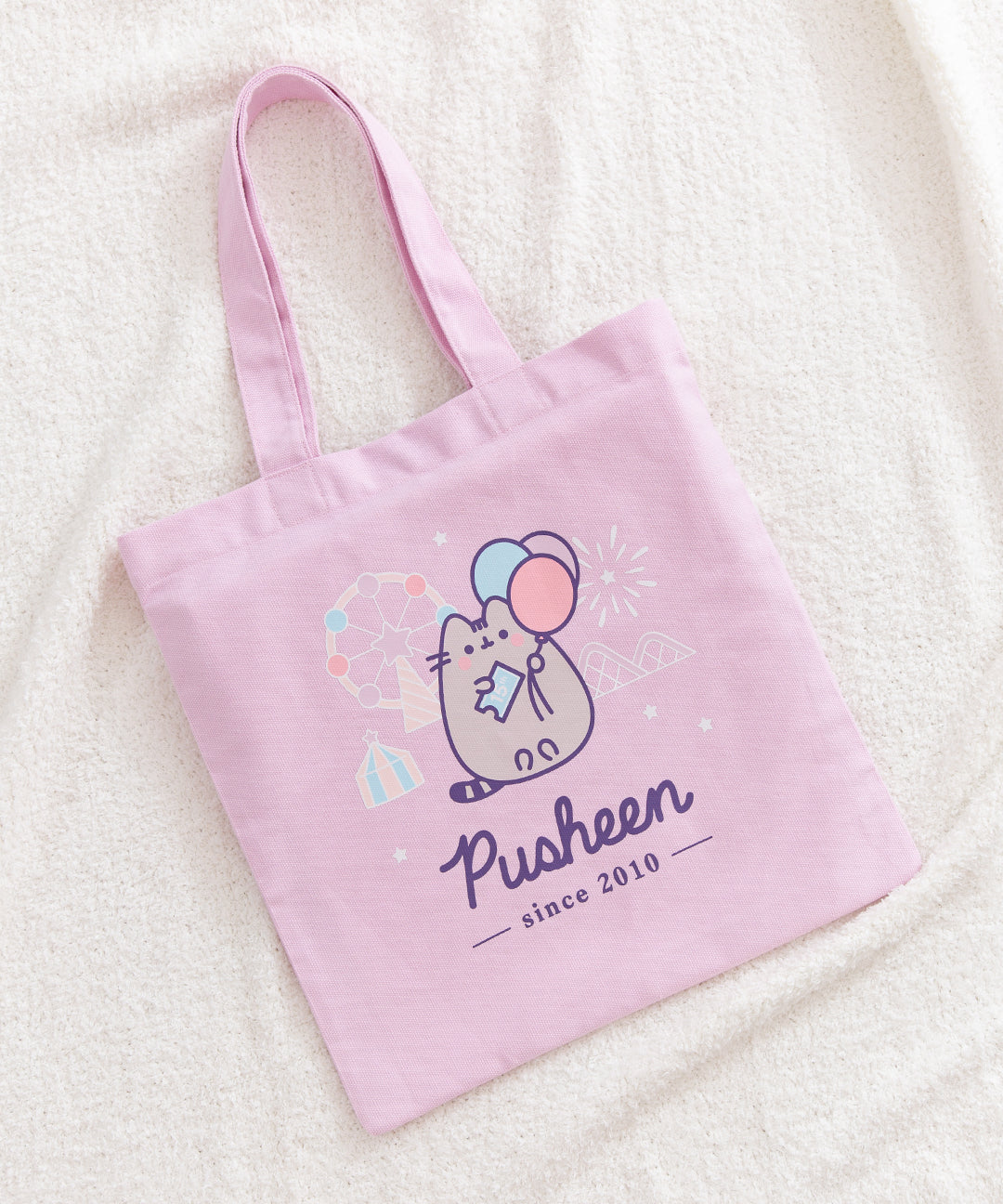 Front view of the Pusheen 15th Anniversary Tote Bag. The large graphic of Pusheen and a carnival take up most of the front of the purple canvas tote bag.