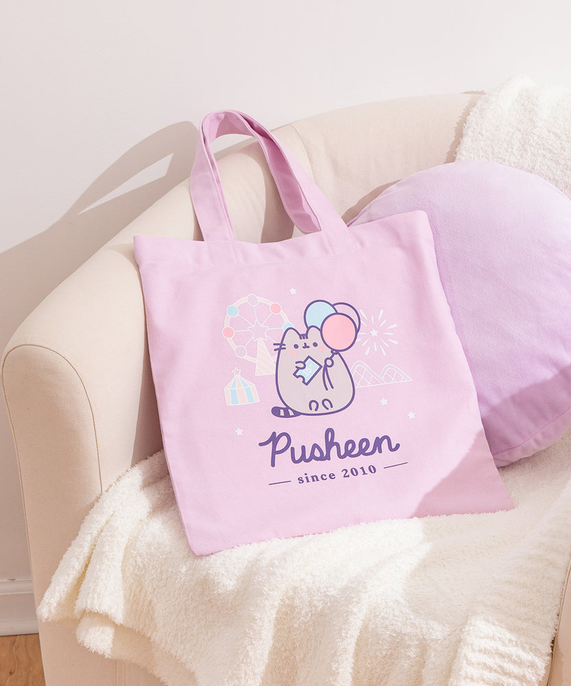 Pusheen 15th Anniversary Tote Bag lies on white chair. The pink canvas tote bag features a Pusheen the Cat holding a ticket to attend a carnival. Below the fair  graphic are the phrases “Pusheen”  and "since 2010."