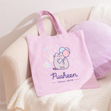 Pusheen 15th Anniversary Tote Bag lies on white chair. The pink canvas tote bag features a Pusheen the Cat holding a ticket to attend a carnival. Below the fair  graphic are the phrases “Pusheen”  and "since 2010."