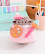 Artist Pusheen Plush in front various art supplies. Pusheen the Cat is dressed to create with a pink beret and smock while holding a paint palette and large paint brush. 