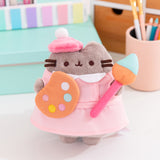 Artist Pusheen Plush in front various art supplies. Pusheen the Cat is dressed to create with a pink beret and smock while holding a paint palette and large paint brush. 