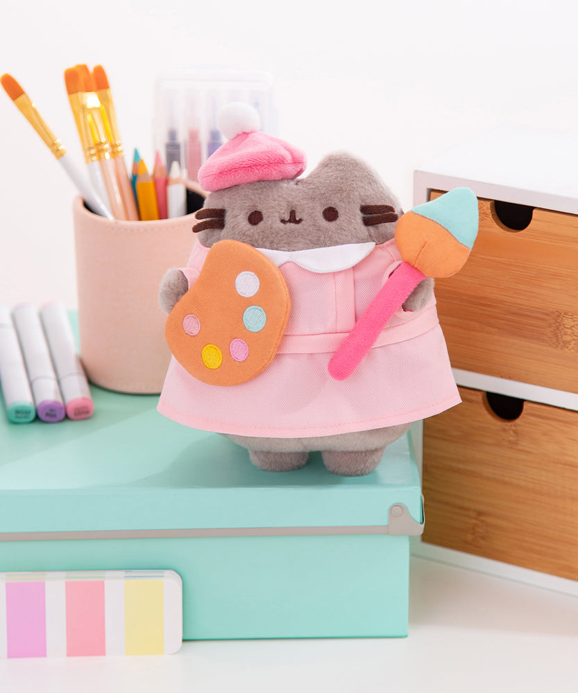 Artist Pusheen Plush standing on two legs in front of various art supplies. The grey fuzzy cat holds a pink paintbrush with blue paint on the bristles.