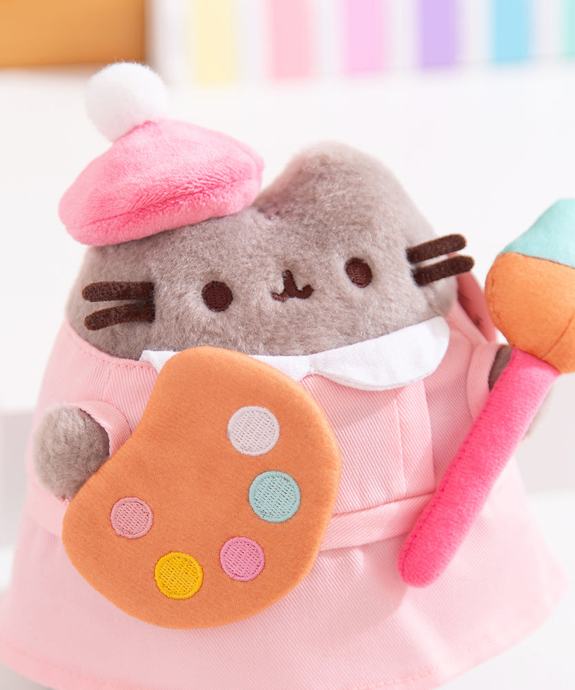 Close-up of Pusheen wearing a fuzzy pink beret. The grey cat has a fluffy texture with brown embroidered facial details. The paint palette that the toy plush is holding has white, light blue, light pink, dark pink, and yellow paint splotches on the brown palette. 