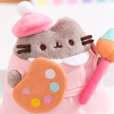 Close-up of Pusheen wearing a fuzzy pink beret. The grey cat has a fluffy texture with brown embroidered facial details. The paint palette that the toy plush is holding has white, light blue, light pink, dark pink, and yellow paint splotches on the brown palette. 