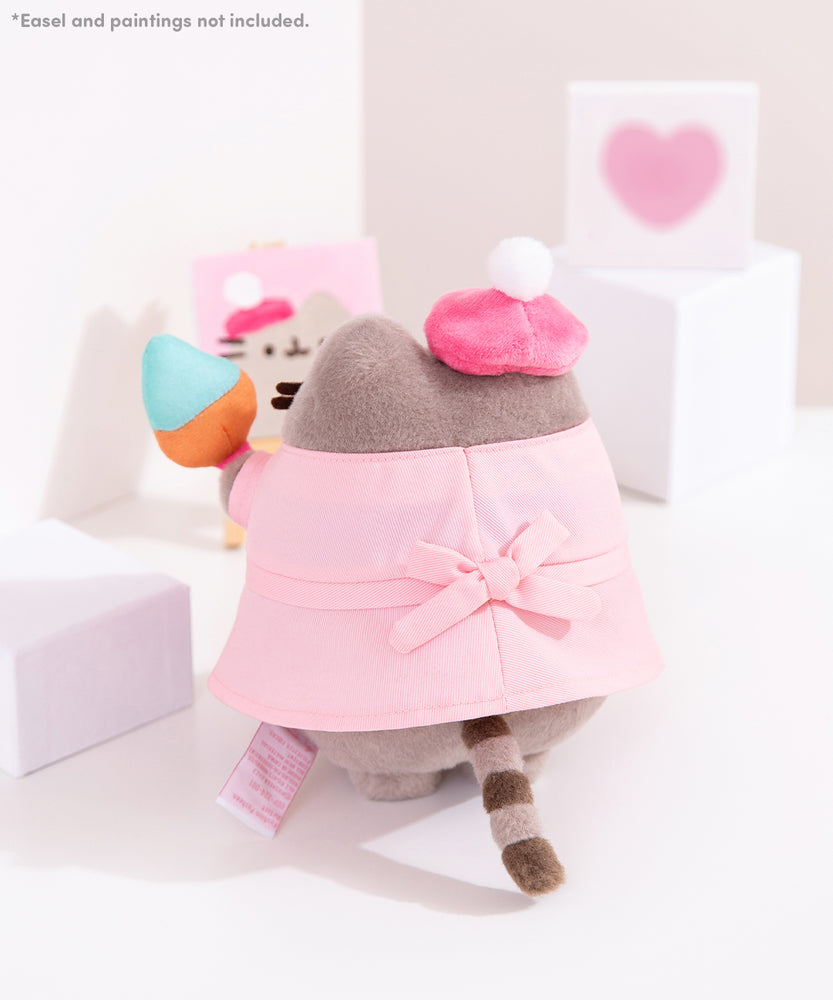 *Easel and paintings not included. Back view of the artist plush. The pink smock has a back bow tie detail. Pusheen's classic striped tail is peeking out from the bottom of the smock.
