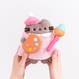 Model holding Pusheen Artist Plush to show size scale. The bottom of the plush fits in the models hands while the head and legs are not encompassed by the hands.