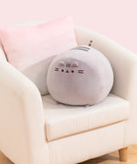 Big Mochi Pusheen Squisheen sits in the corner of a cream chair. The large plush is a light grey color.