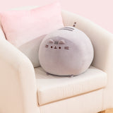 Big Mochi Pusheen Squisheen sits in the corner of a cream chair. The large plush is a light grey color.