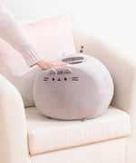 Big Mochi Pusheen Squisheen sits in the corner of a cream chair. A hand pushes down on the top of the grey round plush to show how squishy the plush is.