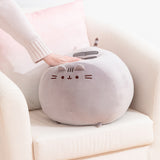 Big Mochi Pusheen Squisheen sits in the corner of a cream chair. A hand pushes down on the top of the grey round plush to show how squishy the plush is.