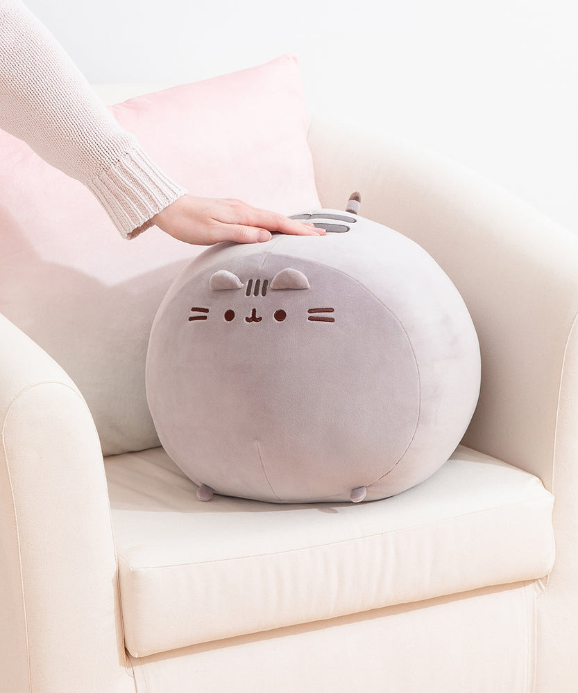 Big Mochi Pusheen Squisheen sits in the corner of a cream chair. A hand rests atop the grey round plush.