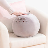 Big Mochi Pusheen Squisheen sits in the corner of a cream chair. A hand rests atop the grey round plush.