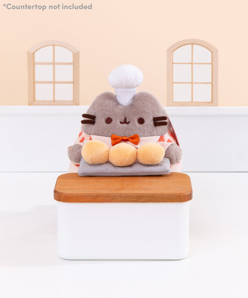 Biscuit Baker Pusheen Plush in front of a light-yellow background. Pusheen holds her three biscuit buns on a platter on top of a wooden countertop. Pusheen’s body is a fuzzy grey material. Image includes the text "*countertop not included" in top left corner of image.