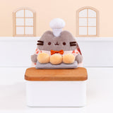 Biscuit Baker Pusheen Plush in front of a light-yellow background. Pusheen holds her three biscuit buns on a platter on top of a wooden countertop. Pusheen’s body is a fuzzy grey material. Image includes the text "*countertop not included" in top left corner of image.