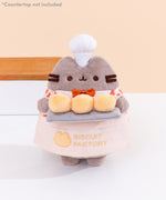 Biscuit Baker Pusheen Plush in front of a light-yellow background. Pusheen the Cat shows off her biscuit baking skills while wearing a checkered shirt with an orange bowtie and an apron that says “Biscuit Factory.” Image includes the text "*countertop not included" in top left corner of image.