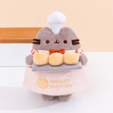 Biscuit Baker Pusheen Plush in front of a light-yellow background. Pusheen the Cat shows off her biscuit baking skills while wearing a checkered shirt with an orange bowtie and an apron that says “Biscuit Factory.” Image includes the text "*countertop not included" in top left corner of image.