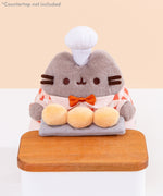 Close-up of Pusheen holds three biscuit buns on a grey slab on a wood countertop. Pusheen wears a white patisserie chef’s hat between her two fuzzy gray ears. Image includes the text "*countertop not included" in top left corner of image.