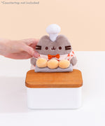 Model holds side of the Baker Pusheen Plush to demonstrate the plush’s size. The checkered shirt Pusheen wears has a light cream, light orange, and burnt orange color pattern. Image includes the text "*countertop not included" in top left corner of image.