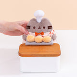 Model holds side of the Baker Pusheen Plush to demonstrate the plush’s size. The checkered shirt Pusheen wears has a light cream, light orange, and burnt orange color pattern. Image includes the text "*countertop not included" in top left corner of image.