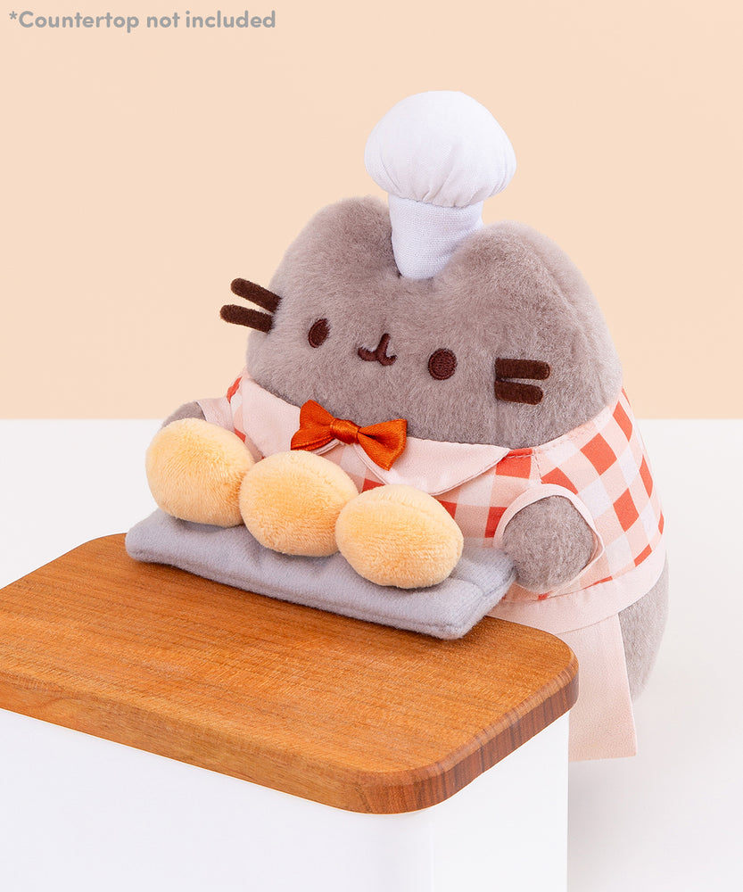 Top aerial view of the Biscuit Baker Pusheen Plush. Pusheen's face is shown in a soft grey plush material and her classic eyes, mouth, and whiskers are shown in dark brown. Image includes the text "*countertop not included" in top left corner of image.