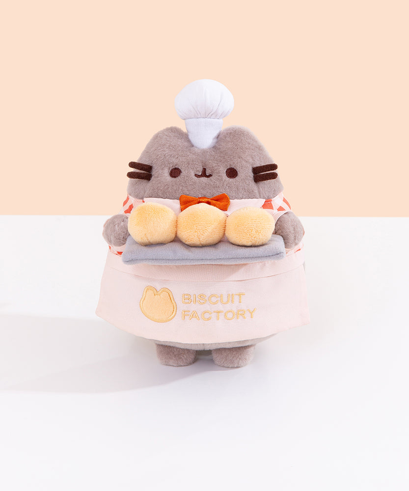 Front view of the Pusheen Baker Plush. Pusheen’s half apron is a light cream color while the embroidered “Biscuit Factory” logo and cat outline are in a light-yellow color that matches the light-yellow color of the biscuits Pusheen holds. 