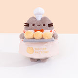 Front view of the Pusheen Baker Plush. Pusheen’s half apron is a light cream color while the embroidered “Biscuit Factory” logo and cat outline are in a light-yellow color that matches the light-yellow color of the biscuits Pusheen holds. 