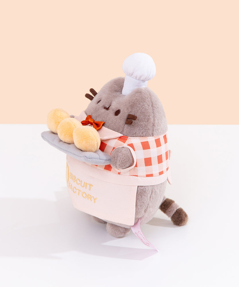 Side view of the Pusheen plush. Pusheen's front paws are holding a tray of biscuits that extends off of the front of the standing plush. 