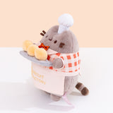 Side view of the Pusheen plush. Pusheen's front paws are holding a tray of biscuits that extends off of the front of the standing plush. 