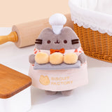   Front view of Biscuit Baker Pusheen Plush. The grey fluffy Pusheen stands in front of a beige rolling pin, whicker bread basket, and a small wooden countertop.