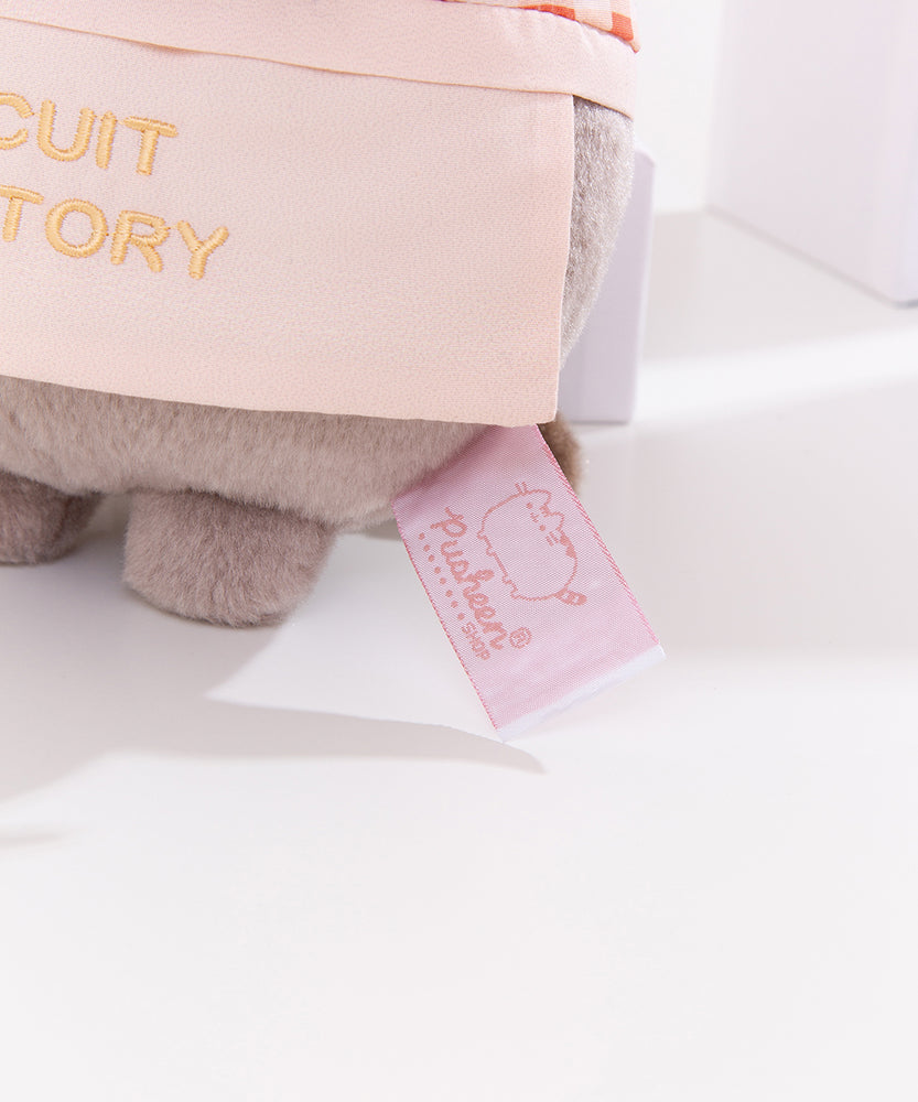 Close-up view of white and pink Pusheen Shop logo tag on the baker plush. 