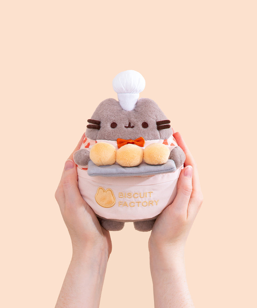 Model holding Biscuit Baker Pusheen Plush to show size scale. The sides of the plush fit in the model’s hands while the head and legs are not encompassed by the hands. 