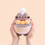 Model holding Biscuit Baker Pusheen Plush to show size scale. The sides of the plush fit in the model’s hands while the head and legs are not encompassed by the hands. 