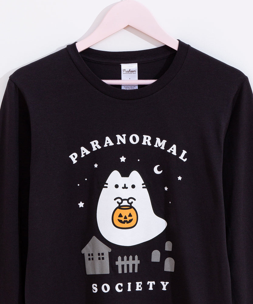 Close-up view of Pusheen Halloween graphic tee. A ghost Pusheen a candy pail while floating in front of a spooky nighttime scene of tombstones, a house, and a fence. Surrounding the ghost are white stars in a variety of sizes and a crescent moon. 