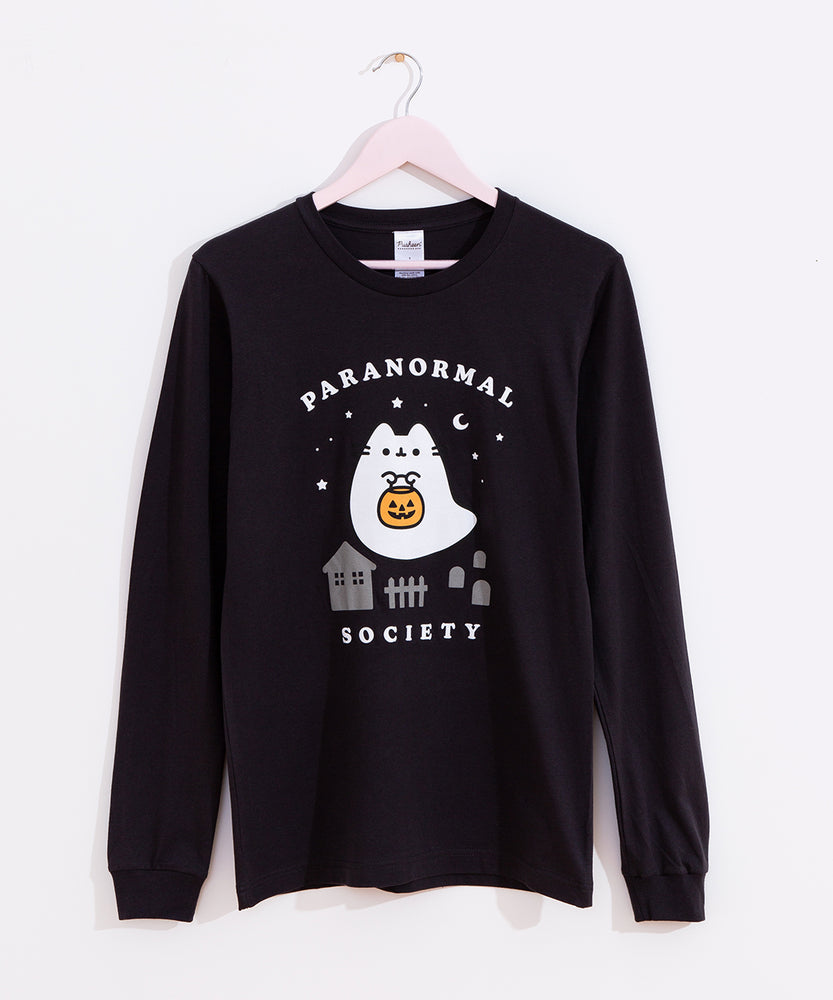 The Boosheen Paranormal Long-Sleeve Unisex Tee hangs on a light pink hanger in front of a white wall. The dark charcoal grey tee shirt features a large graphic of Pusheen the Cat as a friendly ghost that is going trick-or-treating with a jack-o'-lantern candy pail in the front center of the tee. 