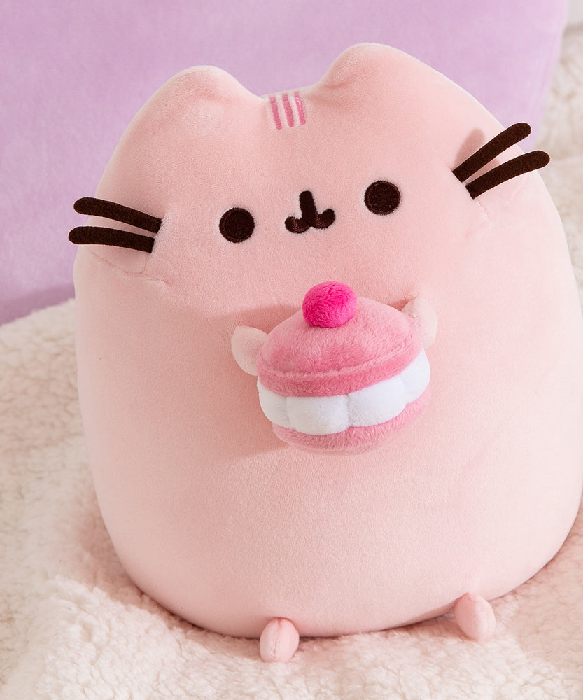 Close-up of the Cherry Squisheen Plush. Pusheen is shown sitting upright and holding a large macaron in her front paws.