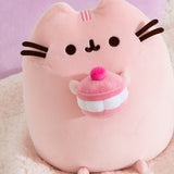 Close-up of the Cherry Squisheen Plush. Pusheen is shown sitting upright and holding a large macaron in her front paws.