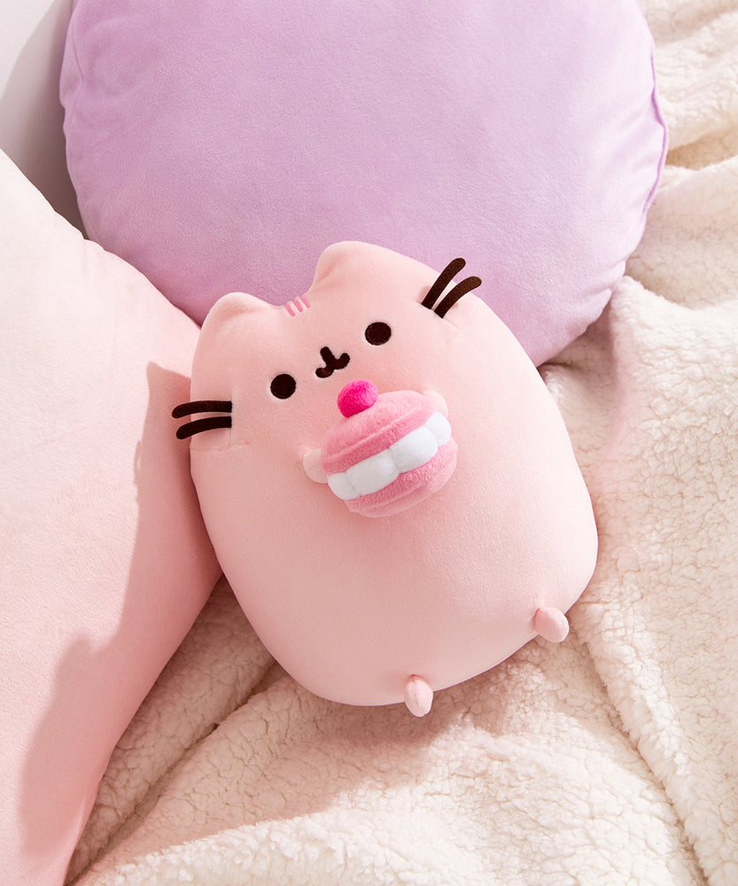 Cherry Plush lies on a white blanket in front of pink and purple pillows. Pusheen's facial features are shown in dark brown.