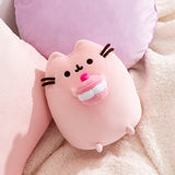 Cherry Plush lies on a white blanket in front of pink and purple pillows. Pusheen's facial features are shown in dark brown.