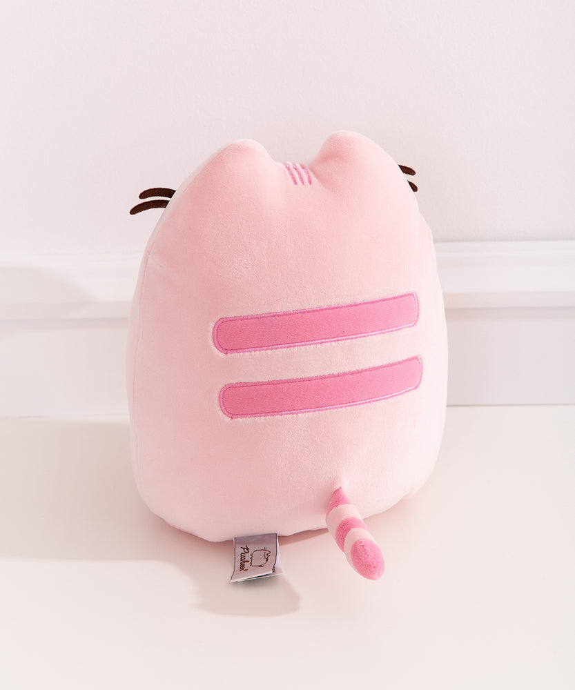 Back view of the Pusheen Sweets Cherry Macaron Squisheen. The light pink plush has two embroidered medium pink stripes along the back. The striped tail extending off the bottom of the plush features the same light pink and medium pink colors alternating.